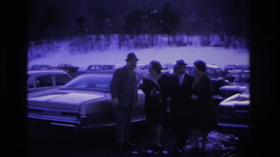Four adults in 1960s winter dress beside a large American sedan in a snowy parking lot; one figure appears to hold an object, with rows of period vehicles and bare trees behind them.