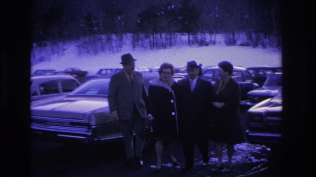 Two couples standing in a snow-covered parking lot in the Hoboken, NJ area, circa 1966; men in fedoras and suits, women in dark winter coats, 1960s sedans behind.