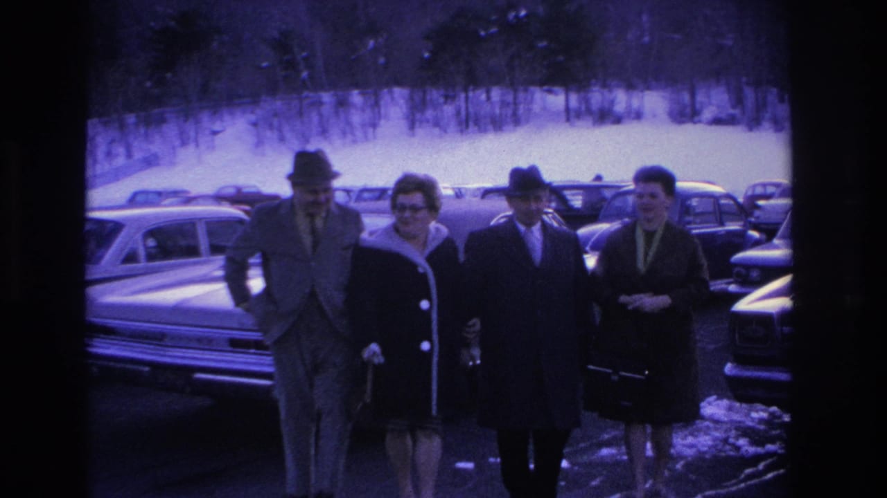 Four adults walking toward camera through a snowy parking lot filled with 1960s cars.