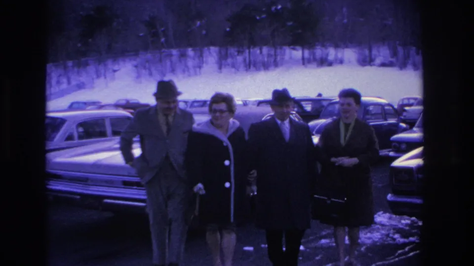 Two couples walking toward camera in a winter parking lot, Hoboken NJ area circa 1966; men in fedoras, women in winter coats, rows of mid-1960s sedans visible.
