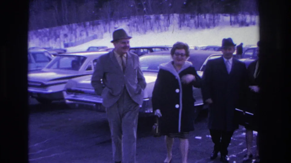 Mid-close view of two couples walking through a winter parking lot, circa 1966; front man in fedora and suit, woman in dark coat with glasses, second couple visible behind.