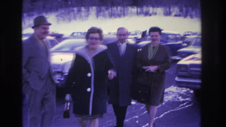 Two couples near camera in a winter parking lot, circa 1966; front man in fedora holds what appears to be a bottle or bag, both women in button-front coats, 1960s sedans behind.