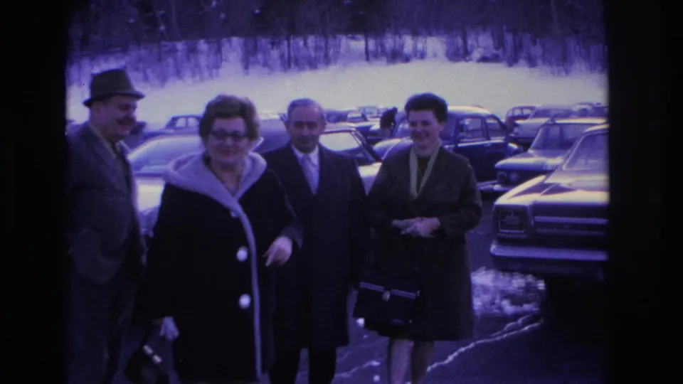 Four adults in 1960s attire, including fedora hats, wool coats, and a fur collar, gather in a parking lot in Hoboken, New Jersey, circa 1966, surrounded by period American sedans.