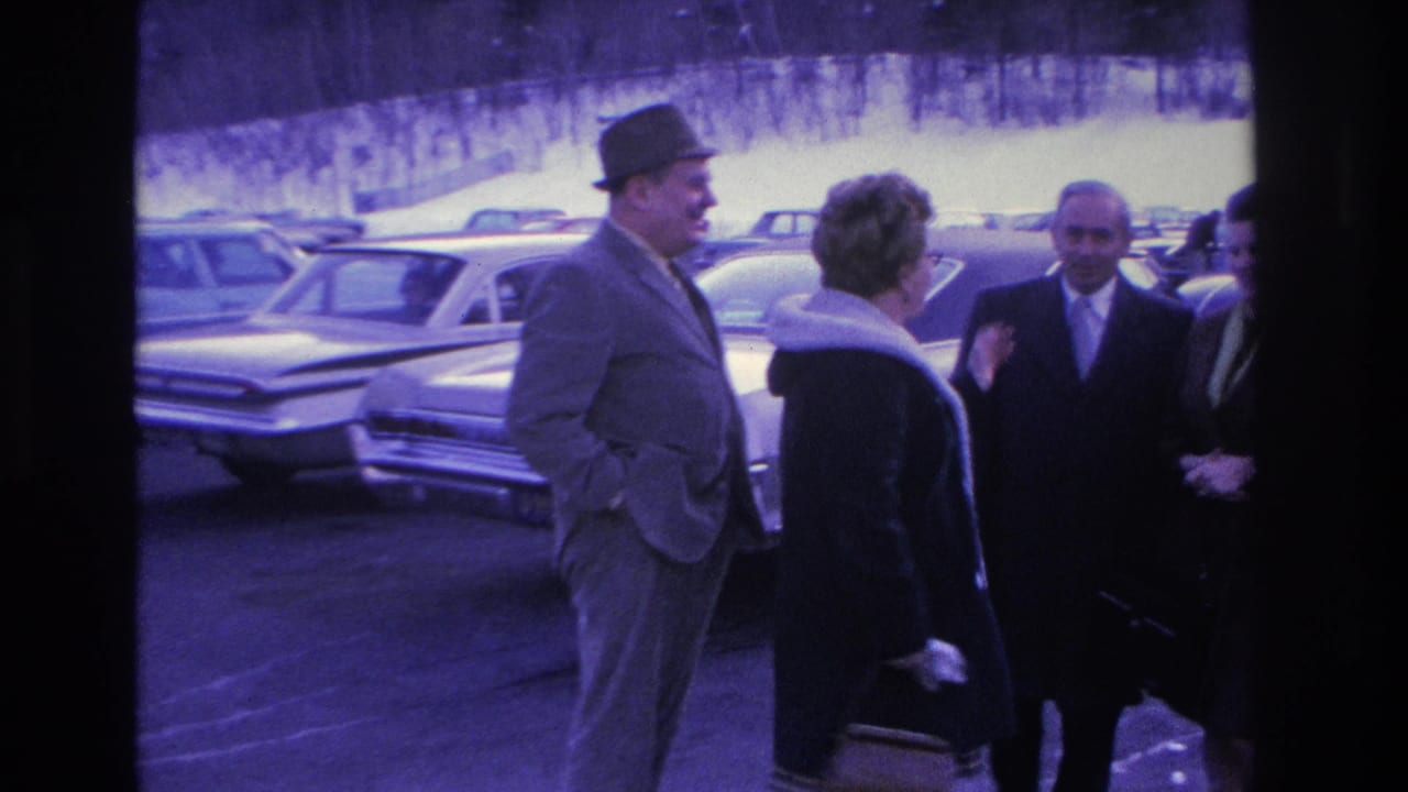 Tall man in fedora and gray suit faces a woman in a dark coat; two other adults and large 1960s sedans visible in background.