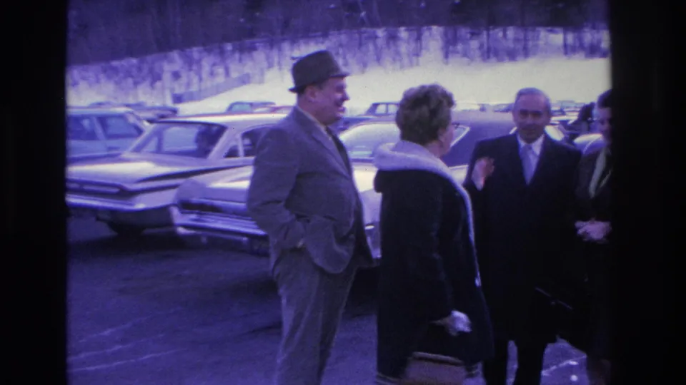 A man in a fedora hat and gray suit engages with a woman in a dark coat in a parking lot, with large early-1960s American automobiles prominent in the background, Hoboken NJ circa 1966.