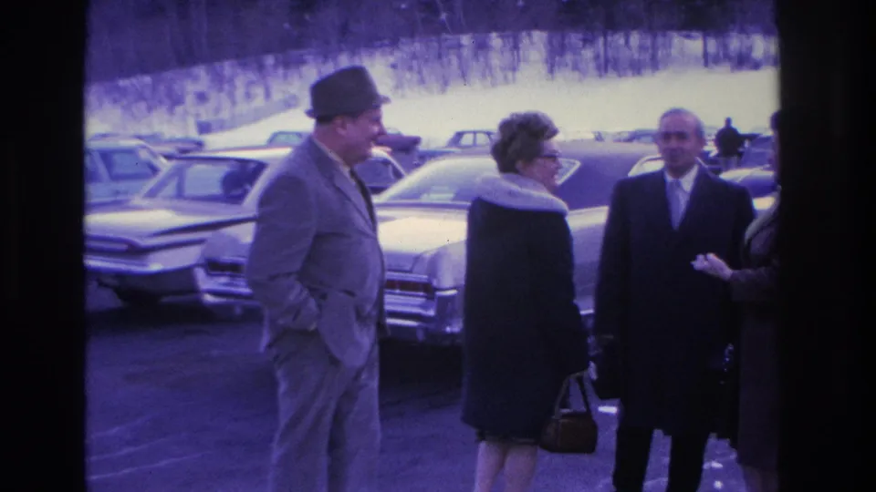 A man in a fedora leans toward a woman in a fur-collared coat and glasses in what appears to be a greeting or kiss, with a third adult and 1960s sedans behind them, Hoboken NJ circa 1966.