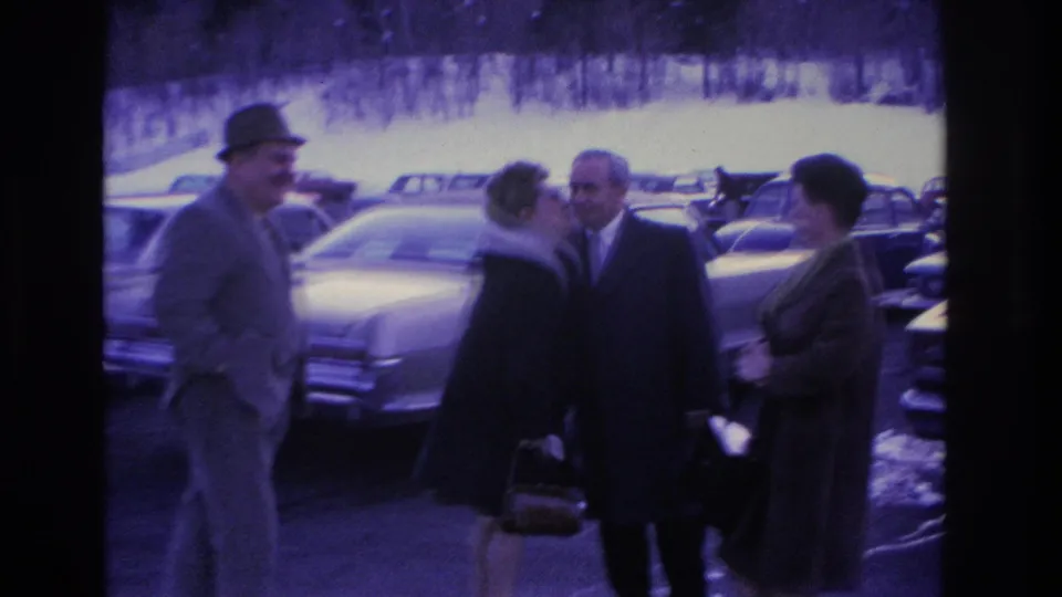 Three adults, a man in a fedora, a woman in a fur-collared coat, and a second man in a suit, share a close greeting moment in a 1960s parking lot, Hoboken NJ circa 1966.