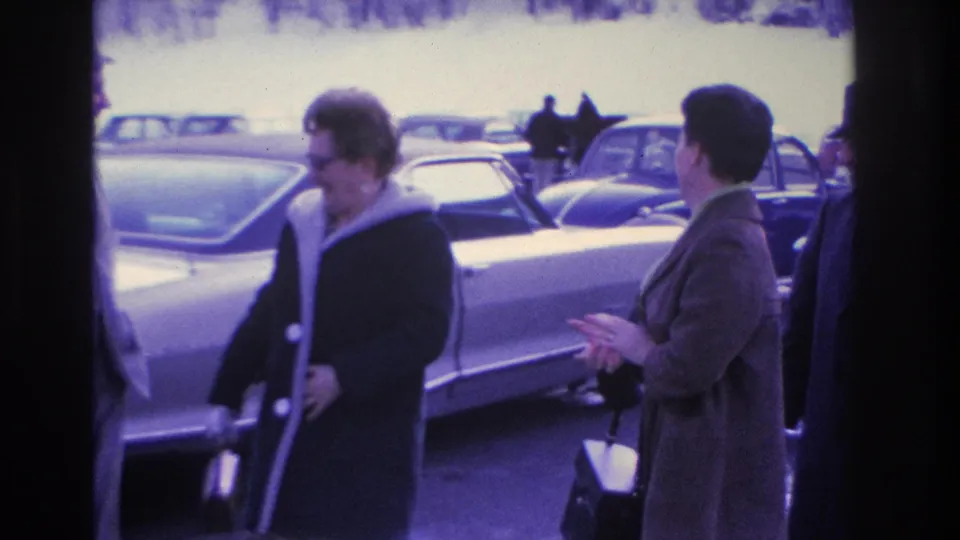 Two women in dark 1960s winter coats, one wearing glasses, gesture animatedly in a parking lot lined with mid-century American cars; circa 1966, Hoboken, New Jersey per metadata.