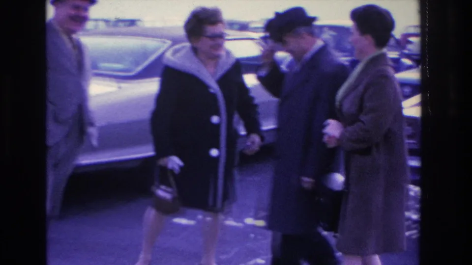 A group of adults, including women in 1960s fur-collar coats and a dark hat, share open laughter in a parking lot; circa 1966, Hoboken, New Jersey per metadata.