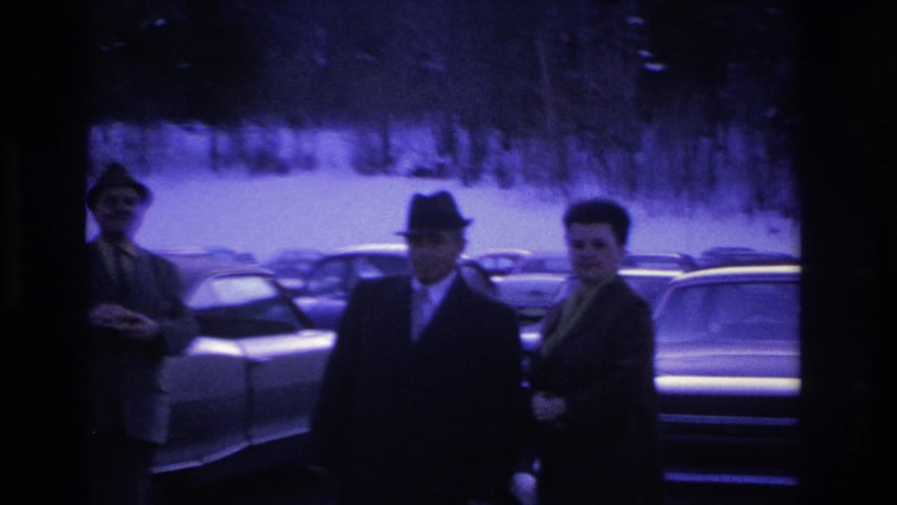 Three suited men in fedoras gathered in a winter parking lot in what appears to be mid-1960s New Jersey, large American sedans visible behind them.