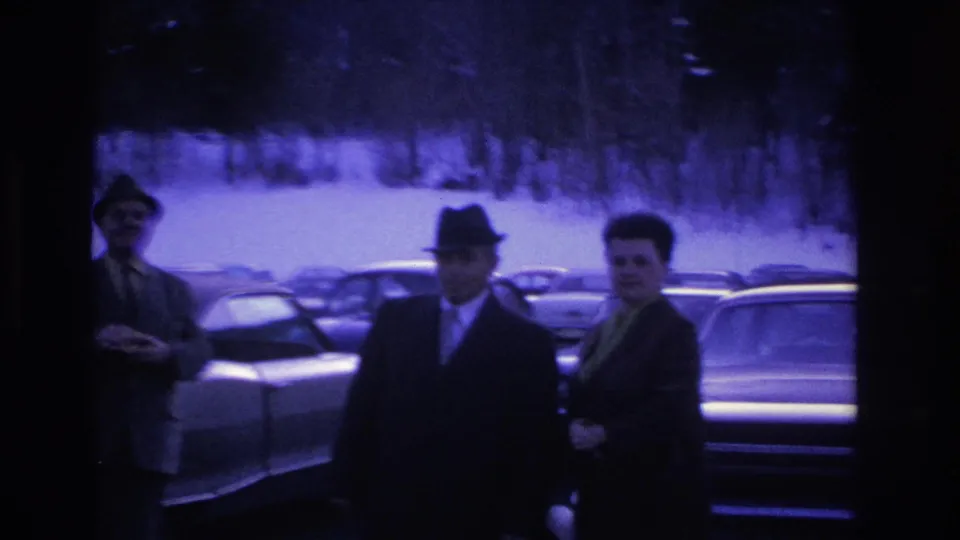 Three suited men in fedoras gathered in a winter parking lot in what appears to be mid-1960s New Jersey, large American sedans visible behind them.