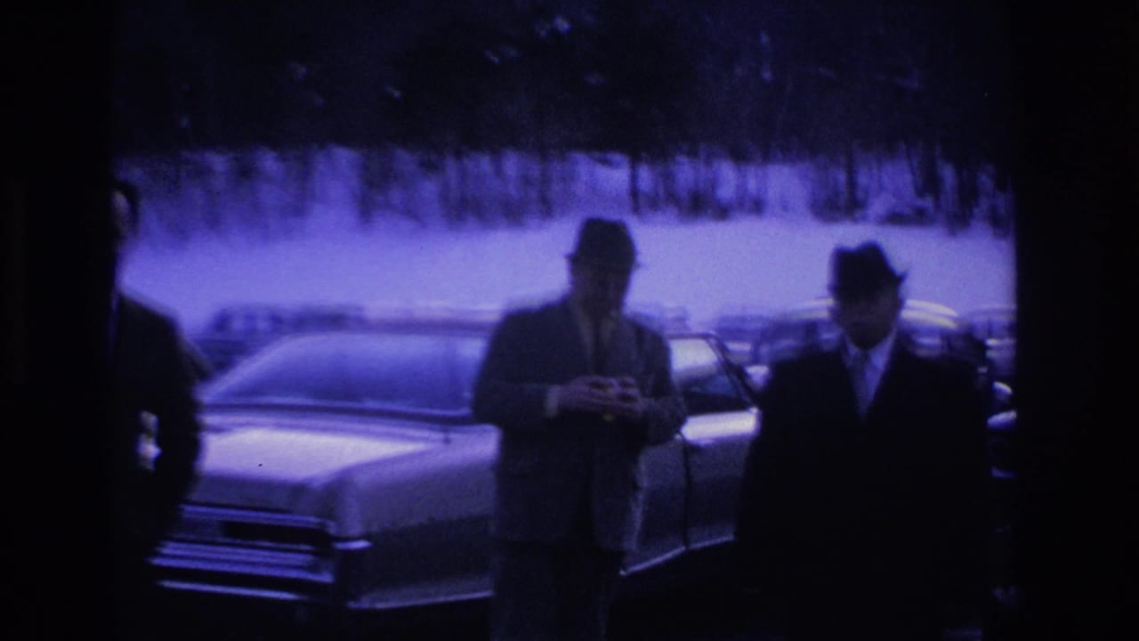 Two men in suits and fedoras stand beside a large 1960s American sedan in a snowy parking lot.