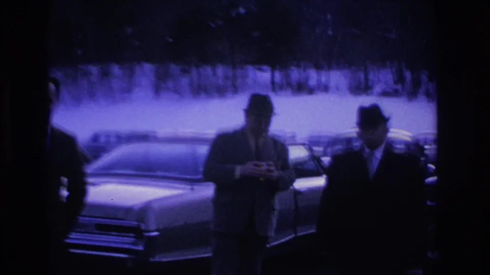 Two suited men wearing fedoras beside a large mid-1960s American sedan in a snow-covered parking lot, captured on 8mm film with a purple color cast.