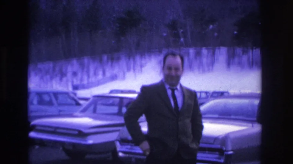 A man in a dark suit and narrow tie laughs directly toward the camera in a winter parking lot, mid-1960s American sedans and snow-covered trees visible behind him.