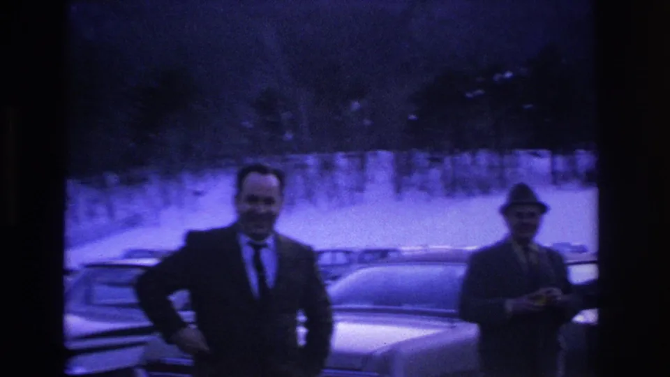 A smiling man in a dark suit and tie poses for the camera while a second hatted man stands behind him in a snow-covered mid-1960s parking lot.