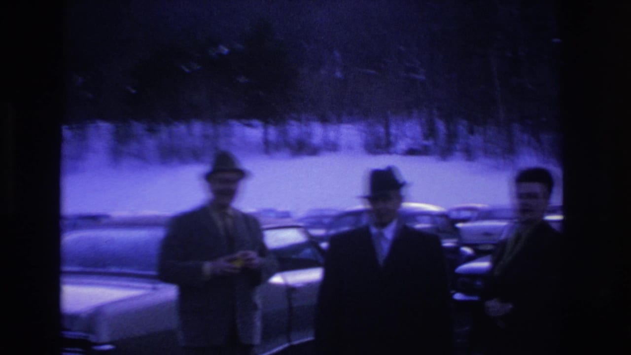 Three men in fedoras and dark overcoats stand in a snow-covered parking lot with mid-1960s American cars; pronounced blue dye-shift on aged 8mm stock.