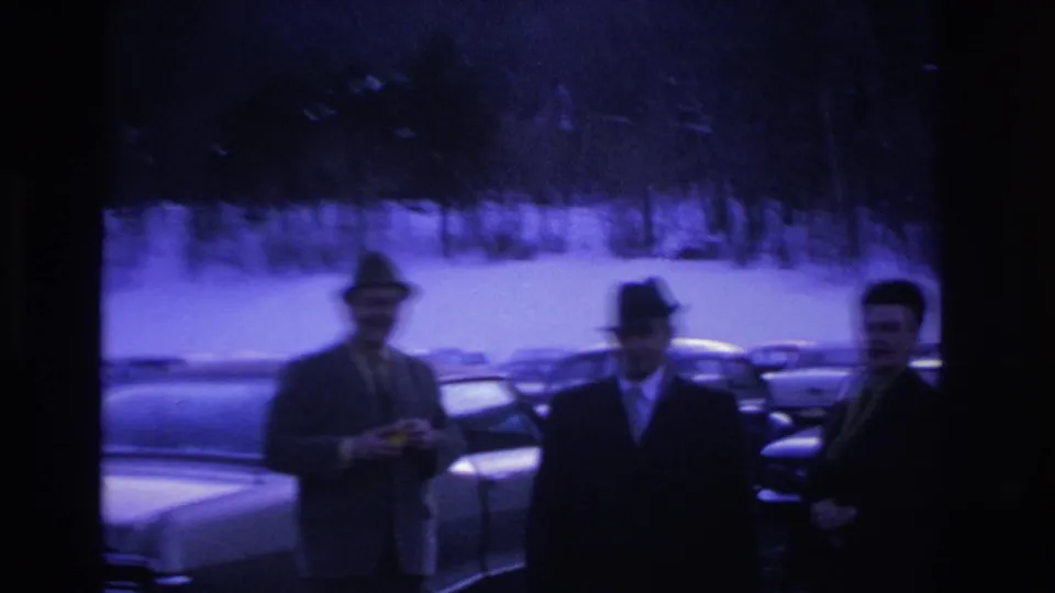 Three men in fedoras and dark overcoats stand in a snow-covered parking lot with mid-1960s American cars; pronounced blue dye-shift on aged 8mm stock.