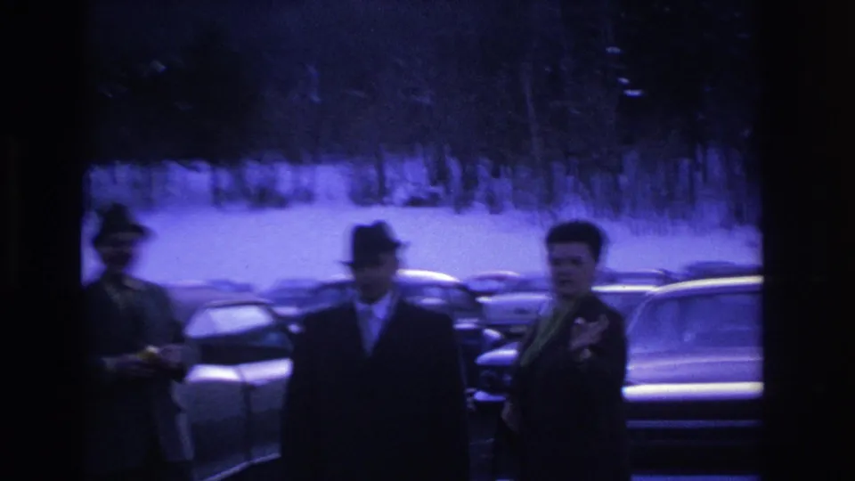 Three men in 1960s-era coats and hats captured mid-movement in a snowy parking lot, with large rounded American sedans visible behind them.