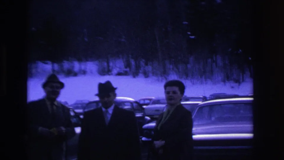 Three men wearing fedoras and dark coats interact casually in a snow-covered parking lot, with 1960s-era American cars and bare trees behind them.