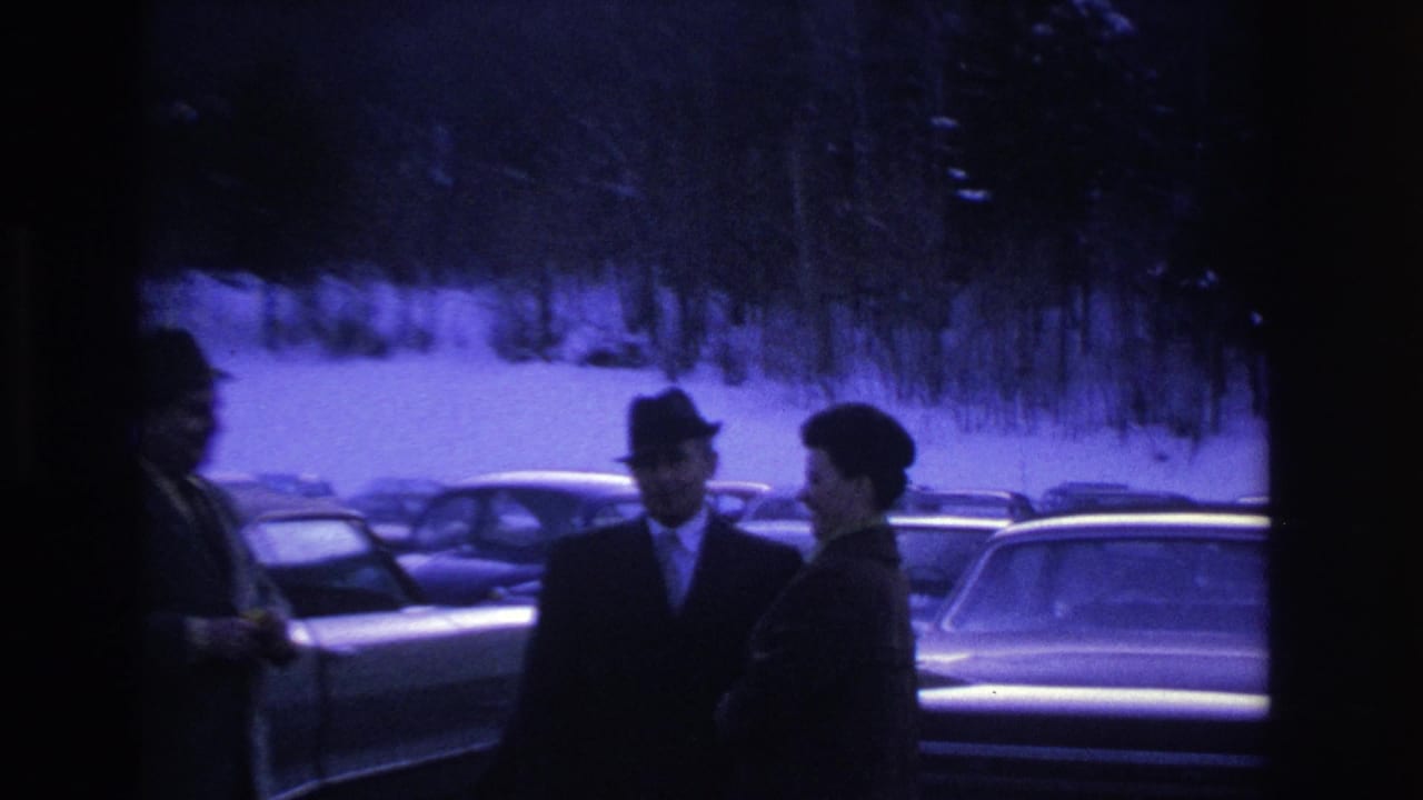 A dark silhouette in the foreground partially obscures two suited men in a snowy parking lot; vintage cars visible behind.
