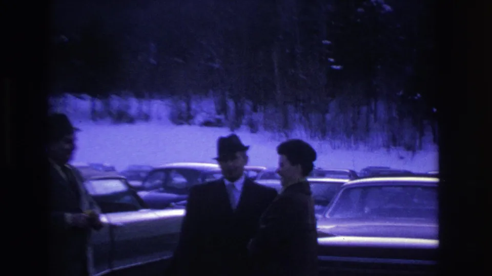 A figure crosses in front of the camera as two men in fedoras and suits remain visible in a snow-covered parking lot; ca. 1966 8mm footage with purple-blue cast.
