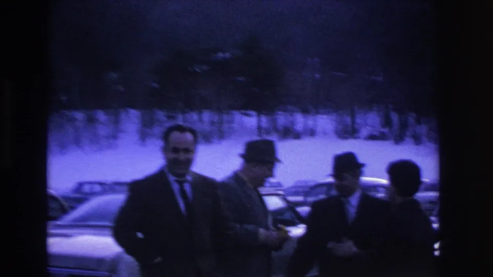 Three to four men in dark overcoats and fedoras grouped in a snowy parking lot, possibly Hoboken NJ area, ca. 1966; vintage American sedans at left edge.