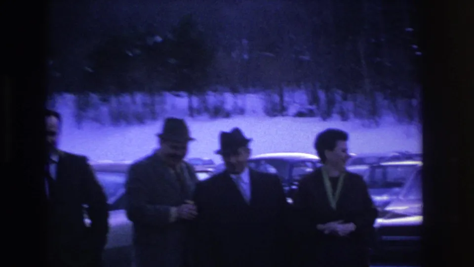 Three men in mid-1960s dress — one in a lighter suit with a colored collar accessory — converse in a snowy parking lot ca. 1966; purple-blue 8mm cast and snow-covered trees behind.