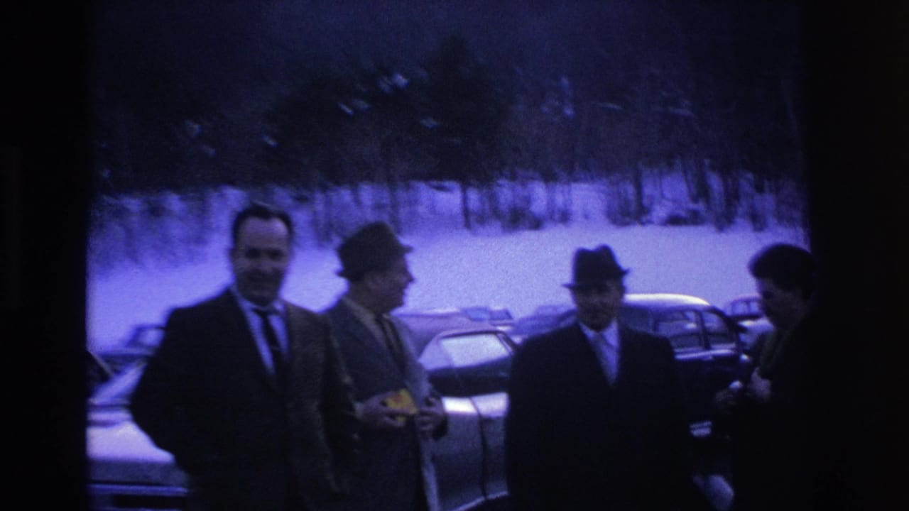 Group of men in suits and fedoras standing in a snowy parking lot, one holding a small object, parked cars and trees behind.