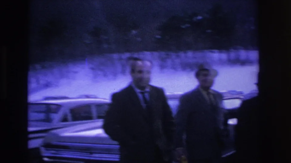 Two men in dark suits walking toward camera in a snowy lot, Hoboken NJ, c.1966; motion blur consistent with handheld 8mm panning.