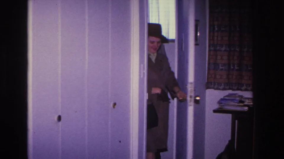Woman in coat and hat stepping through a front doorway into a home interior, c.1966; patterned curtain and side table with papers visible behind her.