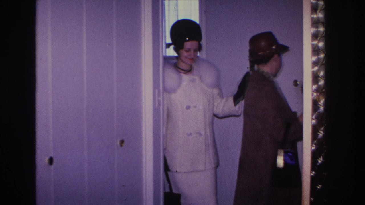 Two adults entering a doorway — woman in white coat and pillbox hat, second figure in brown coat and fedora behind her.