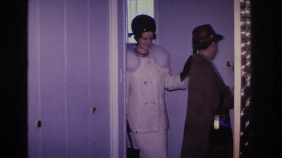 A woman in a white double-breasted coat and black pillbox hat enters a lavender-walled interior with a brown-coated figure behind her; 1966 home movie, Hoboken, New Jersey.