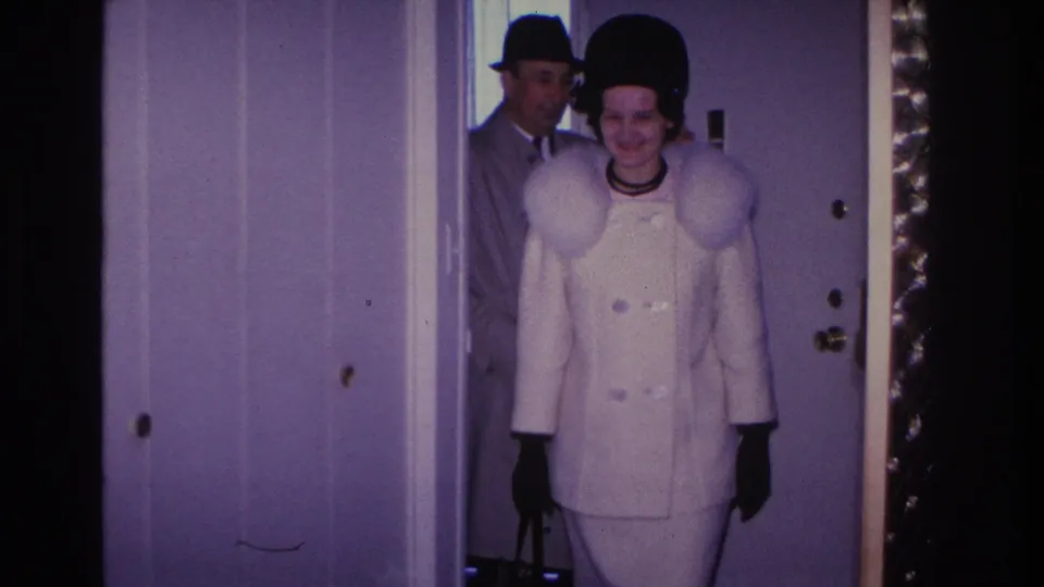 A smiling woman in a white fur-collared coat, black pillbox hat, and dark gloves poses at the doorway of a lavender-walled home; 1966 home movie, Hoboken, New Jersey.
