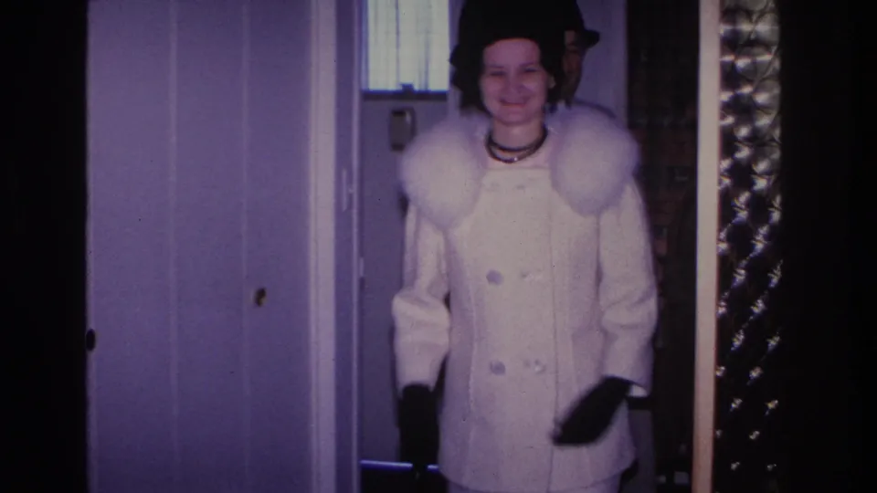 Close view of a woman in a white fur-collared double-breasted coat and black hat smiling inside a mid-1960s home; holiday tinsel garland visible at the door frame.