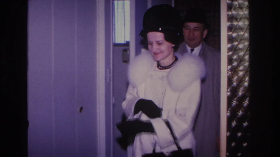 Smiling woman in white fur-collared coat and black hat arriving at a residential doorway, Hoboken New Jersey, circa 1966, 8mm home movie.