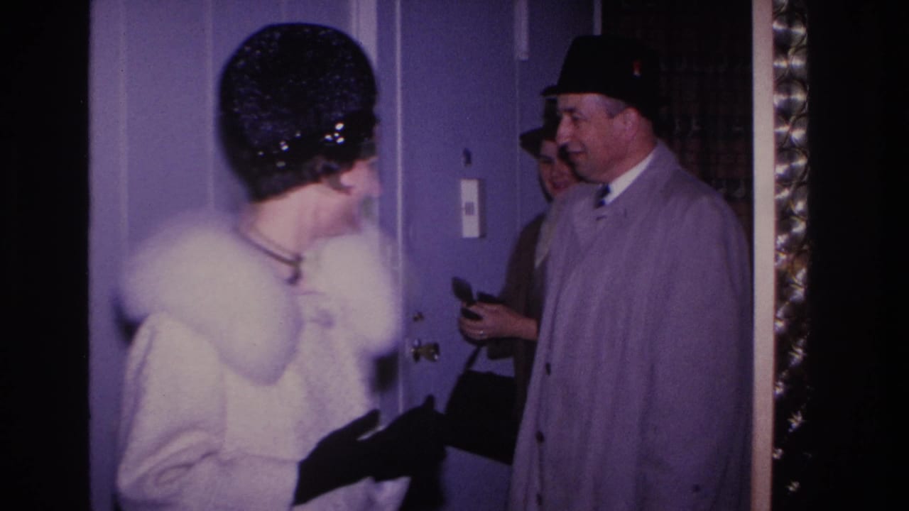 Two visitors at a residential doorway: woman in white fur-collared coat seen from behind, man in gray overcoat holding a small item, circa 1966 8mm home movie.