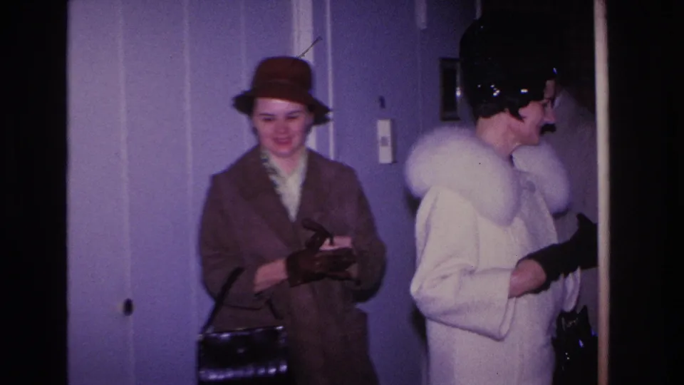Two women inside a residential hallway: one in brown coat and tan hat with a small handbag, one in white fur collar, Hoboken New Jersey, circa 1966.