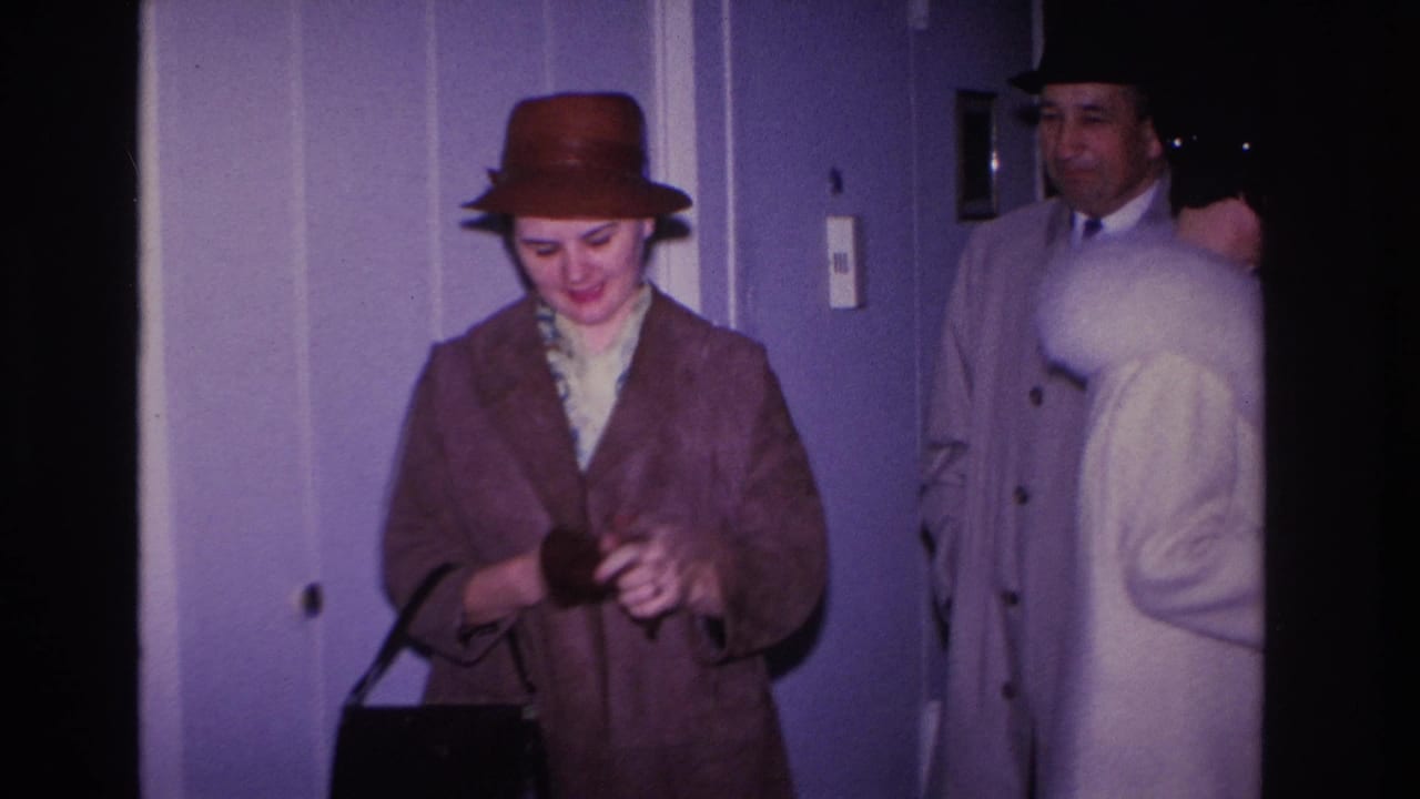 Woman in brown dome hat and brown coat holding a dark handbag; man in light gray overcoat and dark fedora visible behind her in a lavender-walled hallway, Hoboken NJ, c. 1966.