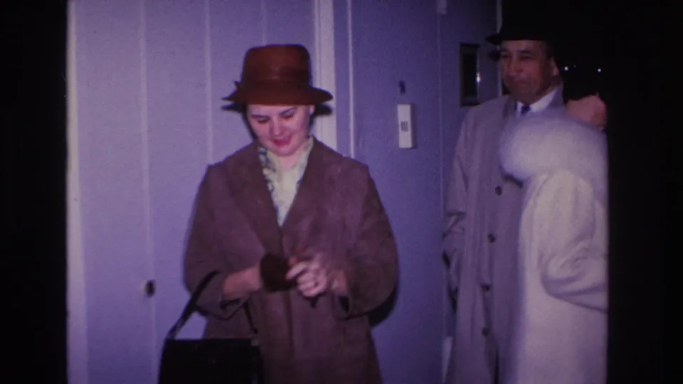 Woman in brown dome hat and brown coat holding a dark handbag; man in light gray overcoat and dark fedora visible behind her in a lavender-walled hallway, Hoboken NJ, c. 1966.