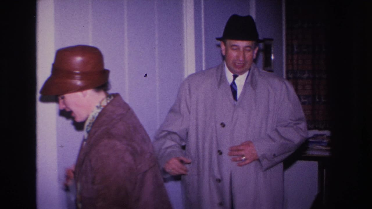 Woman in brown dome hat bows her head near a lavender wall; man in gray overcoat and dark fedora stands beside her, smiling.