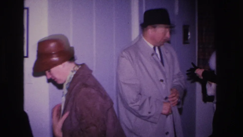 Woman in brown dome hat turns aside while removing her brown coat; black gloves visible; man in gray overcoat and black fedora stands beside her in lavender-walled interior, c. 1966.