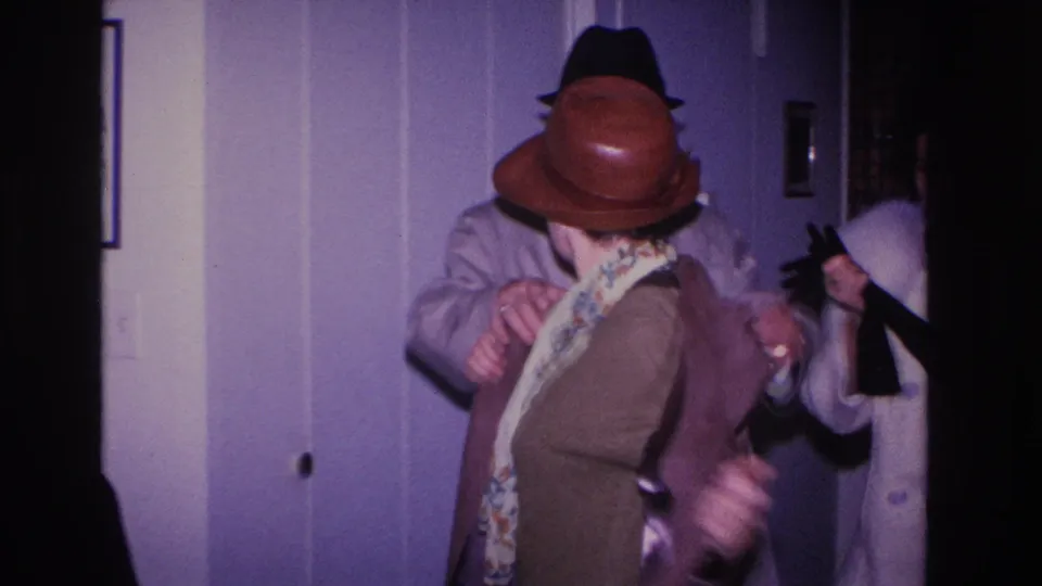 Woman in brown dome hat sheds her coat revealing a patterned scarf or garment; a third person in a light coat appears at right; framed item visible on lavender wall, c. 1966.