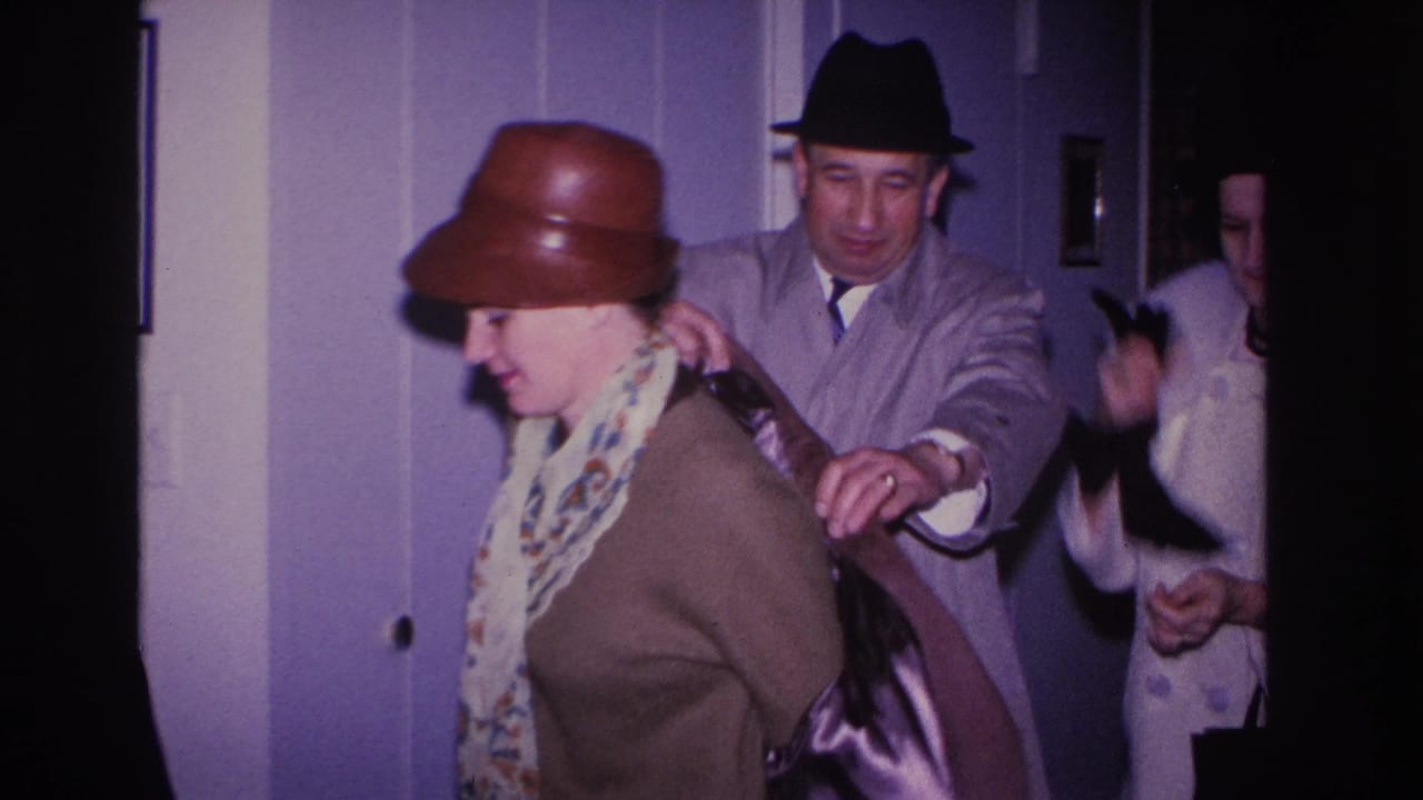 1960s 8mm home movie, Hoboken NJ: man in dark fedora helps woman in brown bowler hat into her coat inside a purple-walled domestic interior.