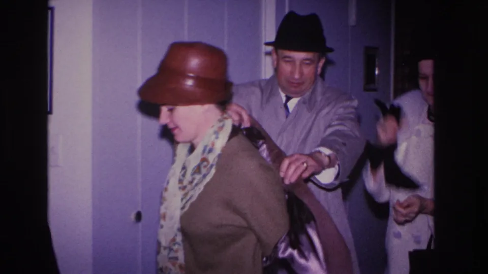 1960s 8mm home movie, Hoboken NJ: man in dark fedora helps woman in brown bowler hat into her coat inside a purple-walled domestic interior.