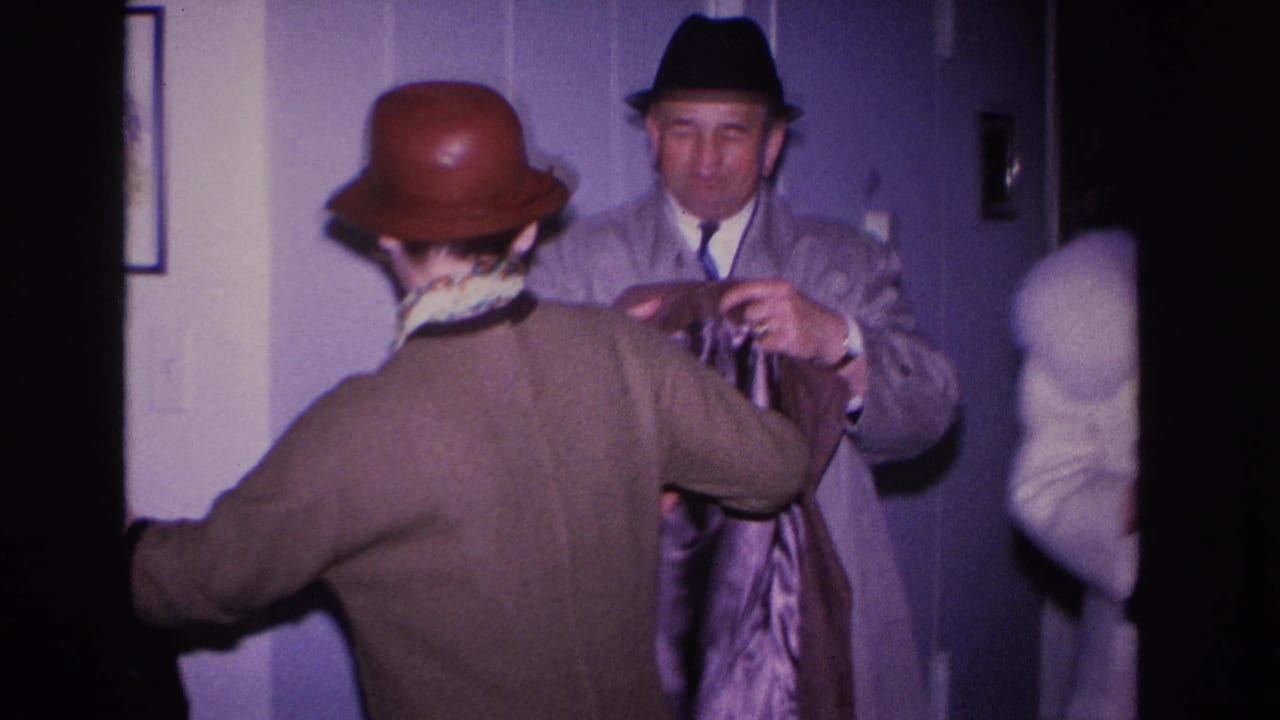 Rear view of woman in brown bowler hat and coat while a man in a gray overcoat and dark fedora adjusts her coat.