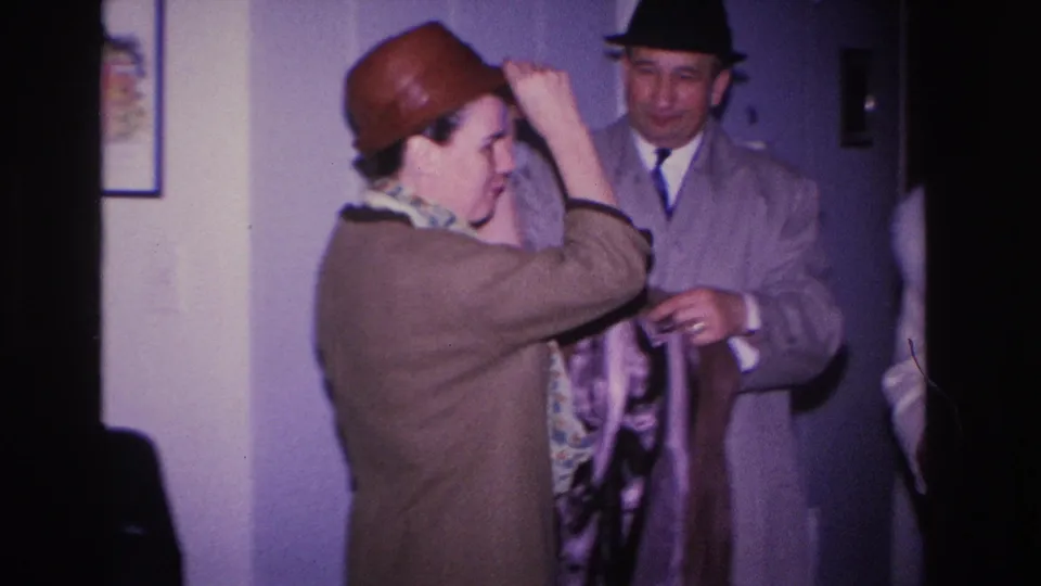 1960s 8mm home movie, Hoboken NJ: woman in brown bowler hat adjusting hat with raised arm, man in dark fedora watching, framed pictures on purple interior wall.