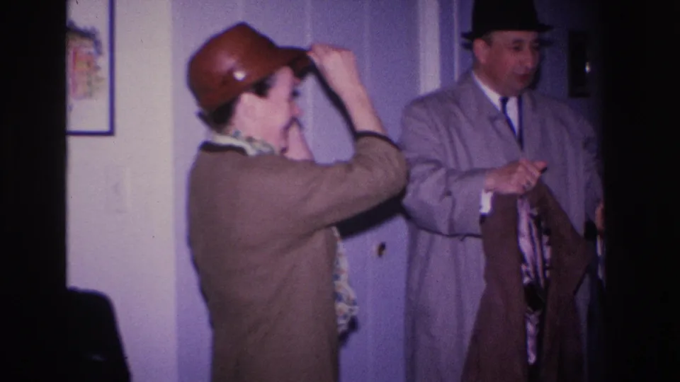 1960s 8mm home movie, Hoboken NJ: woman in brown bowler hat with raised hand adjusting hat, man in dark fedora and gray overcoat alongside, domestic interior with purple walls.