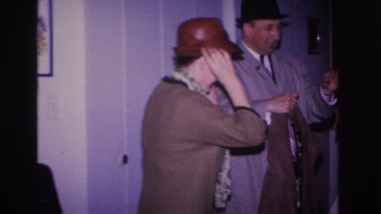 Woman in a brown coat and rounded hat adjusts her hat; man in a gray suit and dark fedora stands beside her, indoor setting Hoboken, New Jersey, circa 1966.