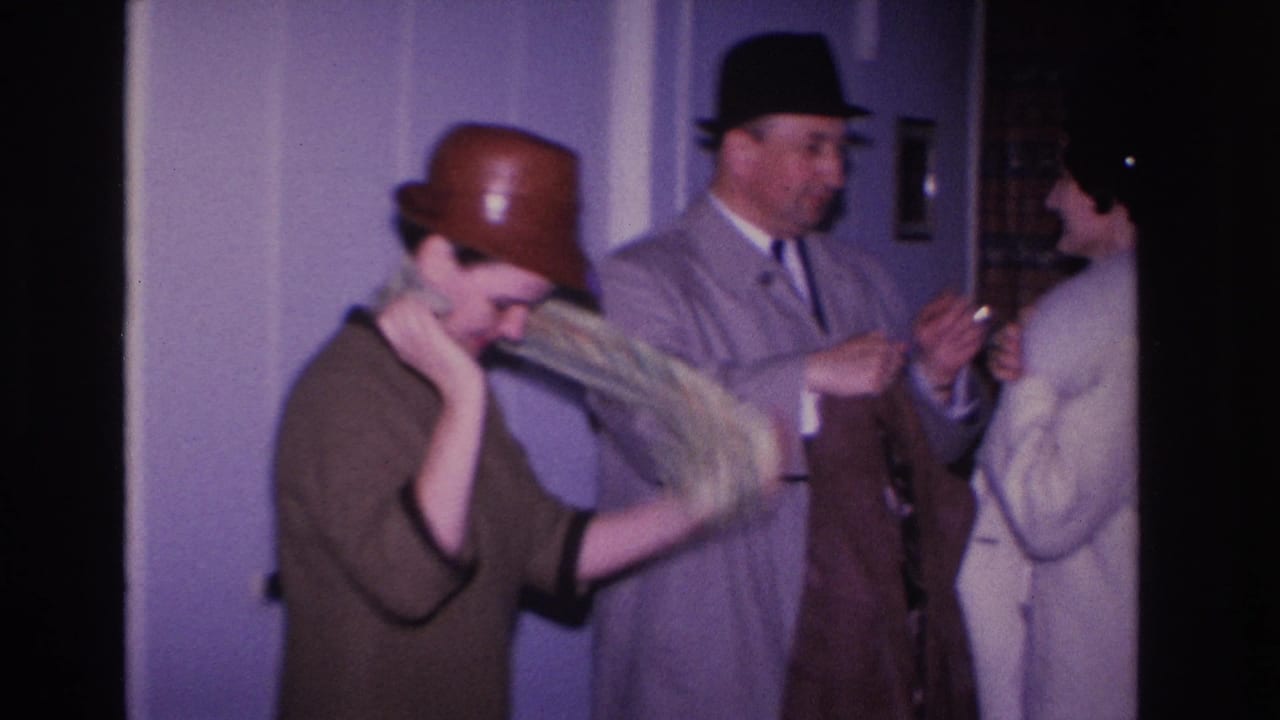 Woman slipping on a coat while a man in a dark fedora stands beside her; a second woman in a light-colored dress is visible at right.