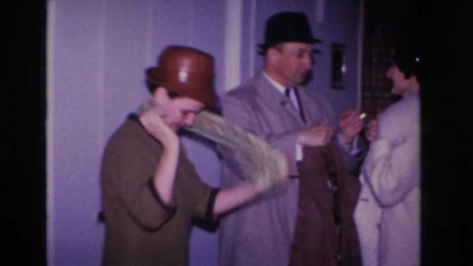 Woman donning a coat with a man in a gray suit and dark fedora at her side and a second woman in a light dress nearby, indoor setting circa 1966.